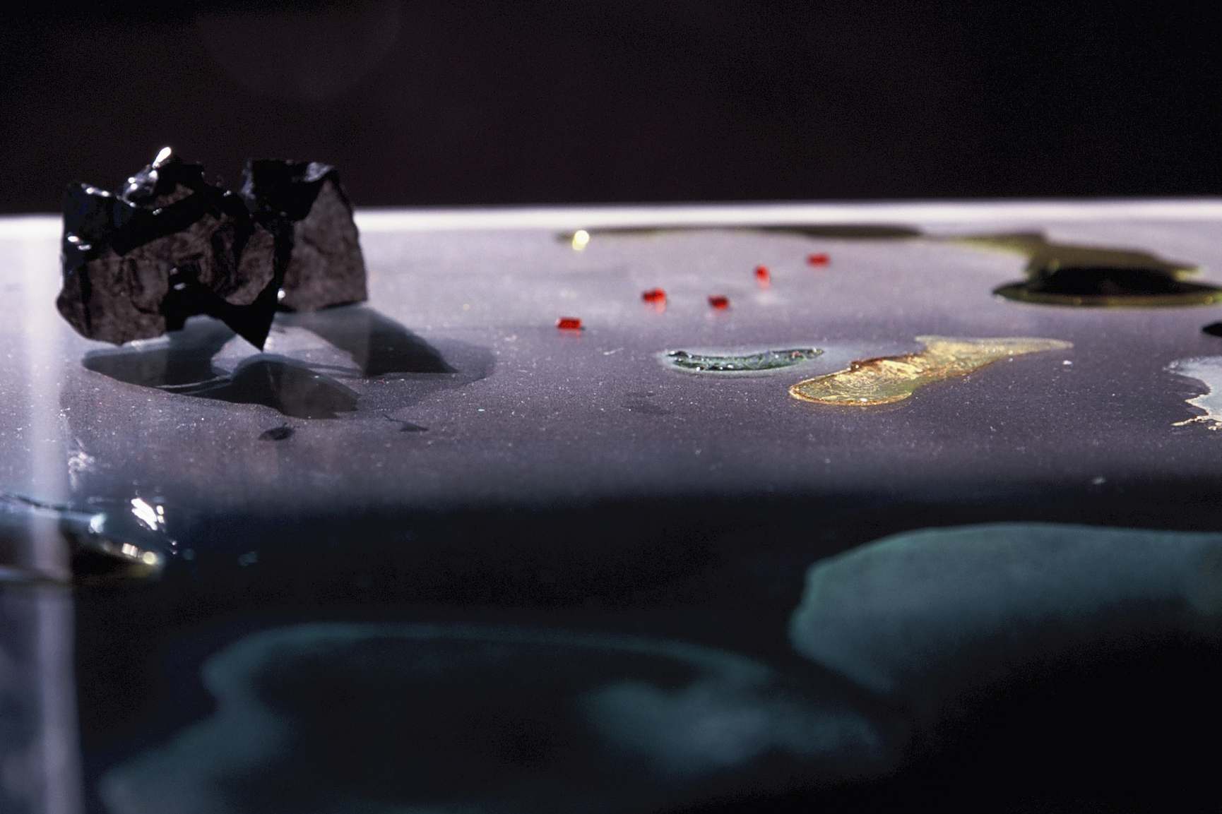 Low-angle close-up of dark solid forms, liquid and red fragments on the glass-topped table